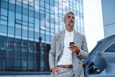 Ein Geschäftsmann hält sein Smartphone in der Hand, während er sein Auto an einer Ladestation für Elektrofahrzeuge auflädt, Nahaufnahme. - HPIF01905