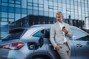 Ein Geschäftsmann hält sein Smartphone in der Hand, während er sein Auto an einer Ladestation für Elektrofahrzeuge auflädt, Nahaufnahme. - HPIF01913