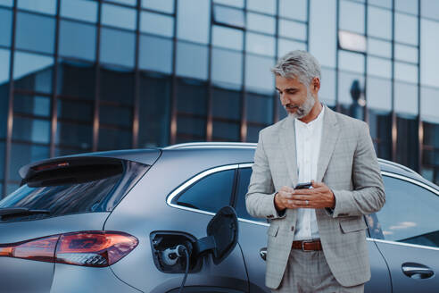 A businessman holding smartphone while charging car at electric vehicle charging station, closeup. - HPIF01915