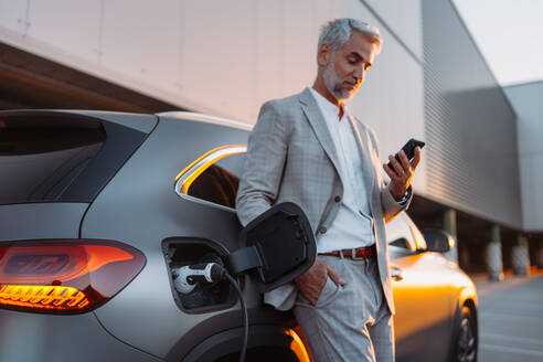 A businessman holding smartphone while charging car at electric vehicle charging station, closeup. - HPIF01934