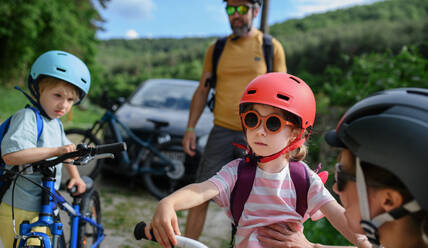 Ein Porträt einer jungen Familie mit kleinen Kindern, die sich auf eine Fahrradtour vorbereiten, mit Fahrrädern und Helm in der Natur. - HPIF01968