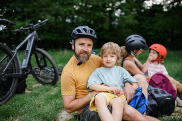 A young family with little children resting after bike ride, sitting on grass in park in summer. - HPIF01987