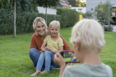 Boy photographing smiling mother and sister through smart phone in garden - JOSEF15018