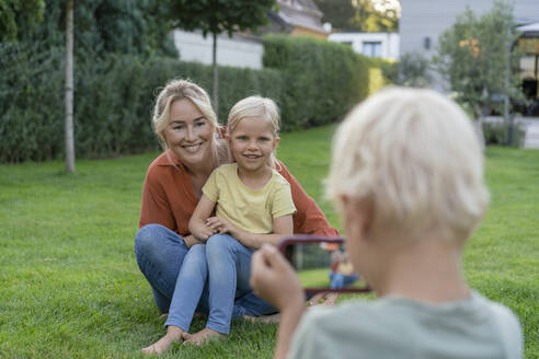 Junge fotografiert lächelnde Mutter und Schwester mit dem Smartphone im Garten - JOSEF15018