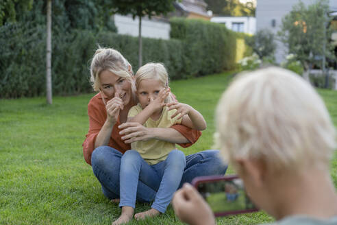 Junge fotografiert Mutter und Schwester, die im Garten mit dem Smartphone gestikulieren - JOSEF15020