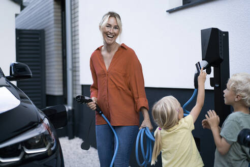 Happy mother holding electric car charger plug by children in front yard - JOSEF15036