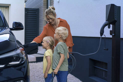 Smiling mother charging electric car by children in front yard - JOSEF15037