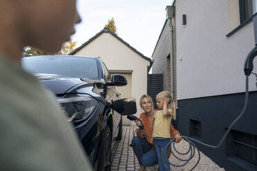 Frau hält den Stecker eines Ladegeräts für Elektroautos und sieht ihre Tochter im Vorgarten an - JOSEF15039