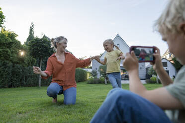 Boy photographing mother dancing with girl through mobile phone in garden - JOSEF15056