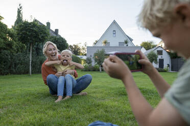 Boy photographing playful sister and mother through mobile phone in garden - JOSEF15059