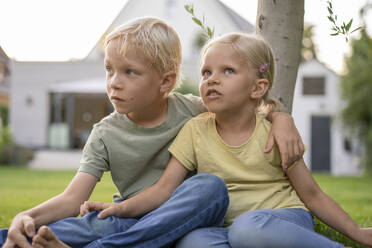 Brother sitting with arm around sister on grass in garden - JOSEF15073