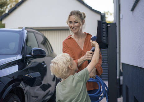 Boy paying through credit card by woman holding electric vehicle charger plug in front yard - JOSEF15084