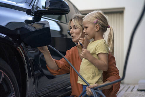 Girl holding credit card by mother charging electric vehicle in front yard - JOSEF15088