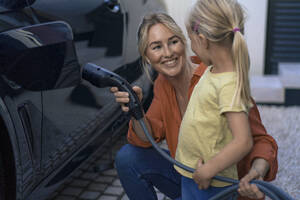 Lächelnde Frau, die den Stecker des Ladegeräts für Elektrofahrzeuge hält und ihre Tochter im Vorgarten betrachtet - JOSEF15091