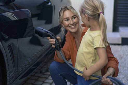 Smiling woman holding electric vehicle charger plug looking at daughter in front yard - JOSEF15091