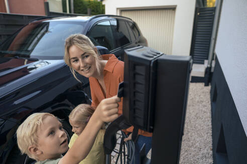 Sohn zahlt mit Kreditkarte, während die Mutter den Stecker des Ladegeräts für Elektrofahrzeuge im Vorgarten hält - JOSEF15097