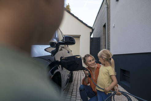 Girl looking at mother holding electric vehicle charger plug in front yard - JOSEF15101