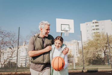 Ein glücklicher Vater und eine Teenager-Tochter, die sich umarmen und in die Kamera schauen, draußen auf dem Basketballplatz. - HPIF02015