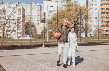 Ein glücklicher Vater und eine Teenager-Tochter, die sich umarmen und in die Kamera schauen, draußen auf dem Basketballplatz. - HPIF02019