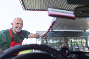 Fröhlicher älterer Angestellter, der an einer Tankstelle ein Autofenster wäscht. - HPIF02158