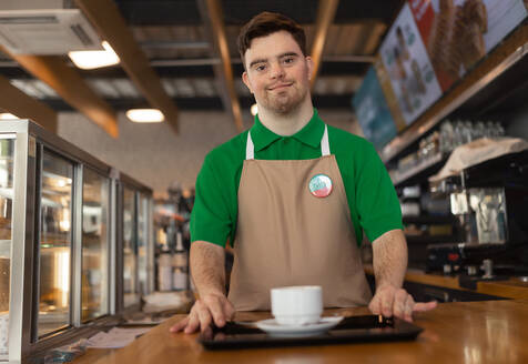Ein glücklicher Kellner mit Down-Syndrom, der einem Kunden in einem Tankstellencafé Kaffee bringt. Konzept der sozialen Integration. - HPIF02175