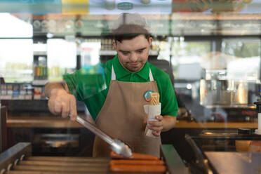 Ein glücklicher Kellner mit Down-Syndrom, der einem Kunden in einem Tankstellen-Café einen Hotdog zubereitet. - HPIF02176