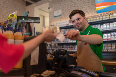 Ein fröhlicher junger Mitarbeiter mit Down-Syndrom, der von einem Kunden in einem Tankstellen-Café Bargeld entgegennimmt. - HPIF02178