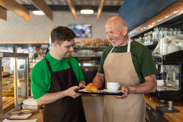 Ein glücklicher Kellner mit Down-Syndrom serviert mit Hilfe seines Kollegen im Café Kaffee. - HPIF02187