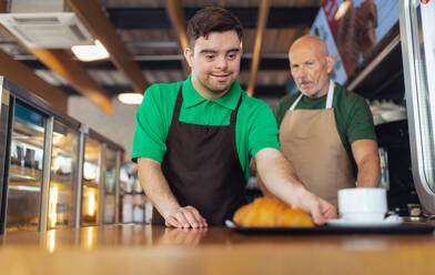 Ein glücklicher Kellner mit Down-Syndrom serviert mit Hilfe seines Kollegen im Café Kaffee. - HPIF02188