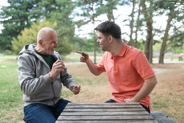 Ein glücklicher älterer Vater mit seinem erwachsenen Sohn mit Down-Syndrom spielt und sitzt im Park. - HPIF02190