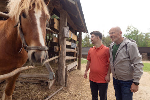 Ein glücklicher älterer Vater mit seinem erwachsenen Sohn mit Down-Syndrom auf einer Ranch, der ein Pferd betrachtet. - HPIF02202