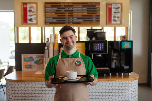 Ein glücklicher Kellner mit Down-Syndrom, der einem Kunden in einem Tankstellencafé Kaffee bringt. Konzept der sozialen Integration. - HPIF02225