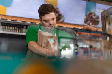 Ein glücklicher Kellner mit Down-Syndrom, der einem Kunden in einem Tankstellen-Café einen Hotdog zubereitet. - HPIF02227