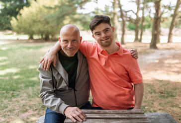 Ein glücklicher älterer Vater und sein kleiner Sohn mit Down-Syndrom umarmen sich und sitzen im Park. - HPIF02234