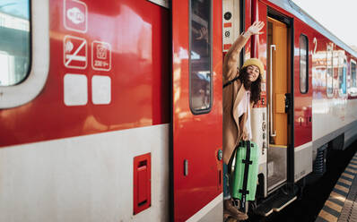 Eine glückliche junge Reisende Frau mit Gepäck steht in der Zugtür und winkt aus dem Zug am Bahnsteig - HPIF02279