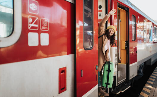 Eine glückliche junge Reisende Frau mit Gepäck steht in der Zugtür und winkt aus dem Zug am Bahnsteig - HPIF02279