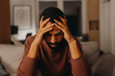 Ein Porträt eines depressiven jungen afroamerikanischen Mannes mit dem Kopf in den Händen, der zu Hause sitzt. - HPIF02393