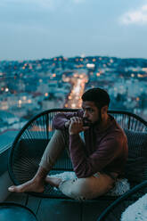 Ein Porträt eines deprimierten jungen afroamerikanischen Mannes, der auf einem Balkon mit Blick auf die Stadt am Abend sitzt. - HPIF02397