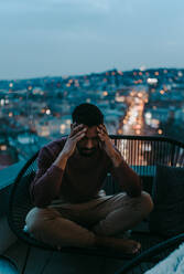 Ein Porträt eines depressiven jungen Afroamerikaners mit dem Kopf in den Händen, der nachts auf einem Balkon sitzt. - HPIF02398