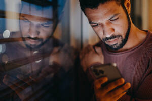 Ein junger Mann mit einem Smartphone schaut traurig, als er abends zu Hause am Fenster steht. - HPIF02400