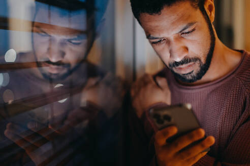 A young man with a smart phone looks sad, standing by window in evening at home. - HPIF02400