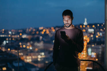 A young man with a smart phone looks sad, standing at balcony with city view in evening at home. - HPIF02401