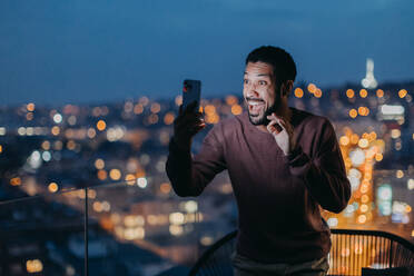 Ein fröhlicher junger Mann macht ein Selfie auf einem Balkon mit Stadtblick. - HPIF02403