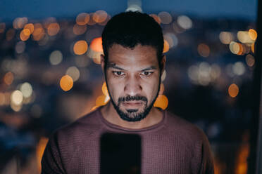 Ein junger Mann mit einem Smartphone sieht besorgt aus, als er abends zu Hause auf dem Balkon mit Blick auf die Stadt steht. - HPIF02404