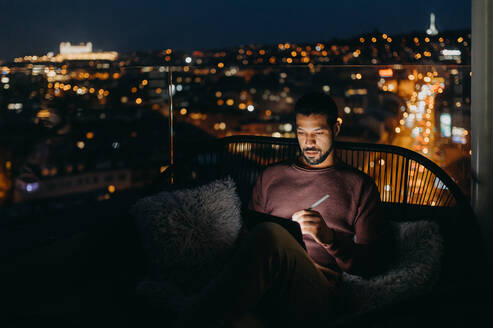 A young African AMerican man sitting on balcony with urban view and using tablet at night - HPIF02406