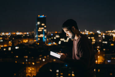 Ein junger afroamerikanischer Mann steht auf einem Balkon mit Blick auf die Stadt und benutzt ein Tablet bei Nacht - HPIF02409