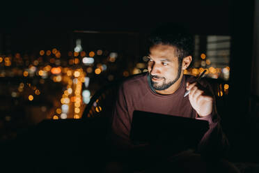 Ein junger afroamerikanischer Mann sitzt auf einem Balkon mit Blick auf die Stadt und benutzt ein Tablet bei Nacht - HPIF02410