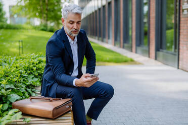 Ein erfolgreicher reifer Geschäftsmann benutzt sein Smartphone, während er sich auf der Straße ausruht, auf einer Bank sitzt und eine mobile Anwendung benutzt. - HPIF02412