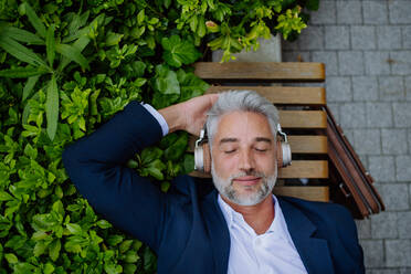 Eine Draufsicht auf einen reifen Geschäftsmann, der auf einer Bank im Stadtpark während einer Arbeitspause Musik mit Kopfhörern genießt. - HPIF02417
