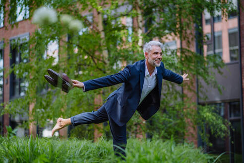 Ein lustiges Porträt eines glücklichen, energischen, reifen Geschäftsmannes, der Schuhe trägt und barfuß läuft und eine Pose im Park einnimmt, sich frei fühlt, Work-Life-Balance-Konzept. - HPIF02427